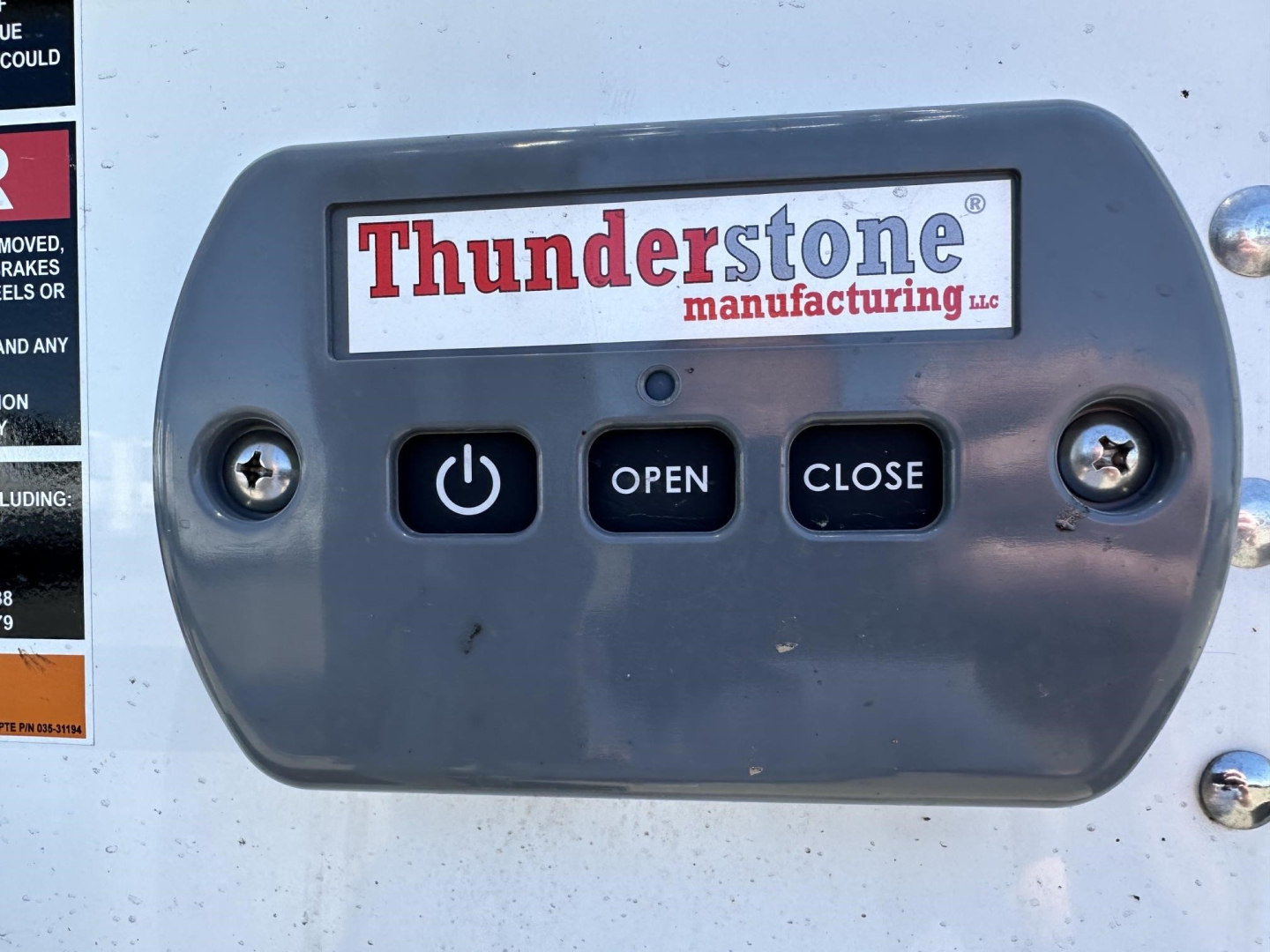 Close-up of a Thunderstone manufacturing power/push button panel with OPEN and CLOSE buttons on a trailer door, with brand name Thunderstone and surrounding screws.