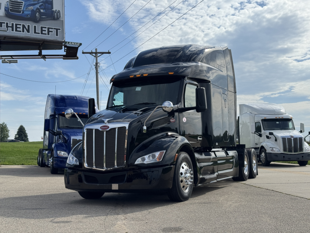 Peterbilt 579 conventional sleeper truck in black, front three-quarter view, with chrome grille and large aerodynamic cab, other trucks visible in background.