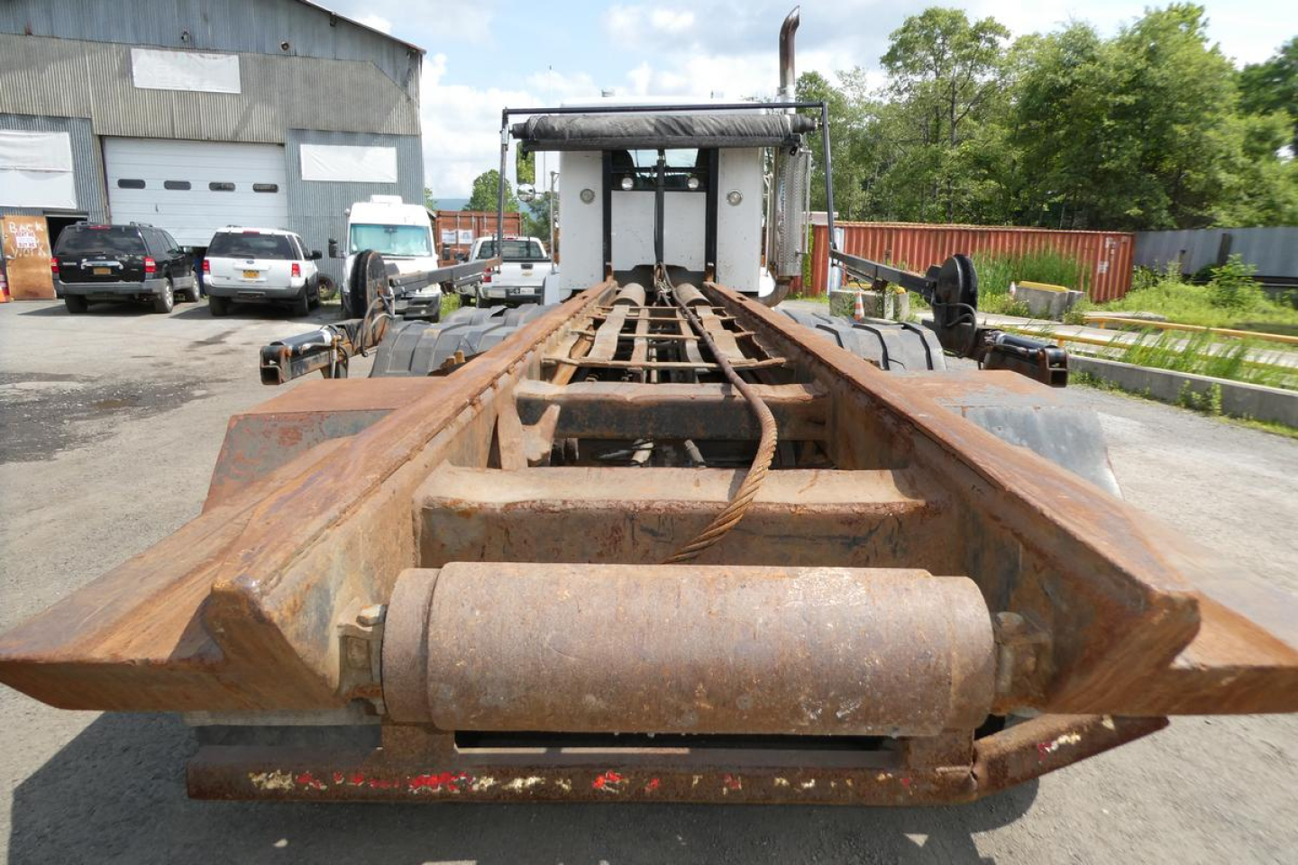 Trailer chassis showing rusted I-beam frame with hitch/platform for rolloff truck, rear view, Kenworth T800 used hull in background.