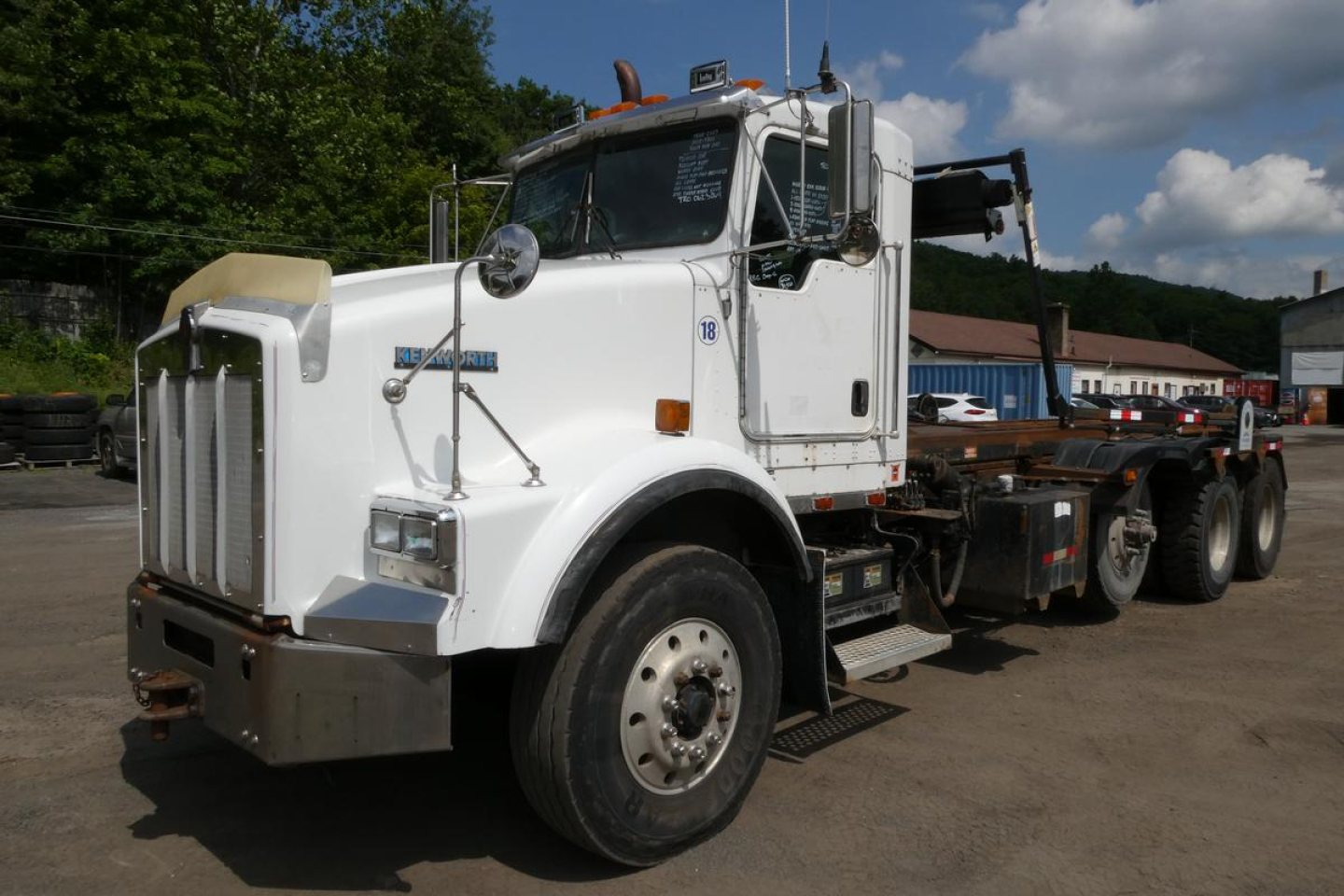 Kenworth T800 roll-off truck in white, side view showing front grille, cab and chassis with hook lift on a sunny day.