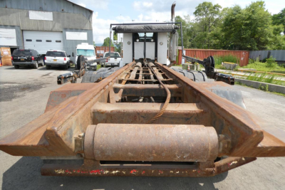 Trailer chassis showing rusted I-beam frame with hitch/platform for rolloff truck, rear view, Kenworth T800 used hull in background.