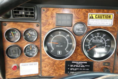 Dashboard panel of a Kenworth T800 showing gauges and warning labels. Visible text includes CAUTION label, Power Take-Off, Hobbs hours gauge, RPM tachometer, and various switches.
