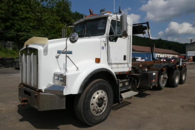 Kenworth T800 roll-off truck in white, side view showing front grille, cab and chassis with hook lift on a sunny day.