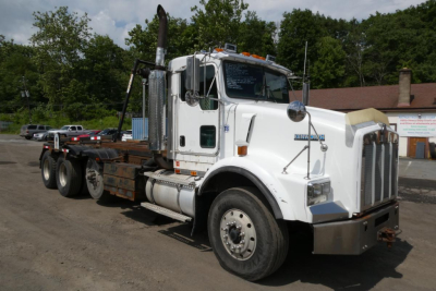 Kenworth T800 rolloff truck in white with flatbed and large exhaust stacks showing VIN window and stock number.