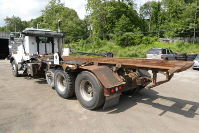 Kenworth T800 rolloff truck rear/side view showing long flatbed with rusted frame and dual rear axles.