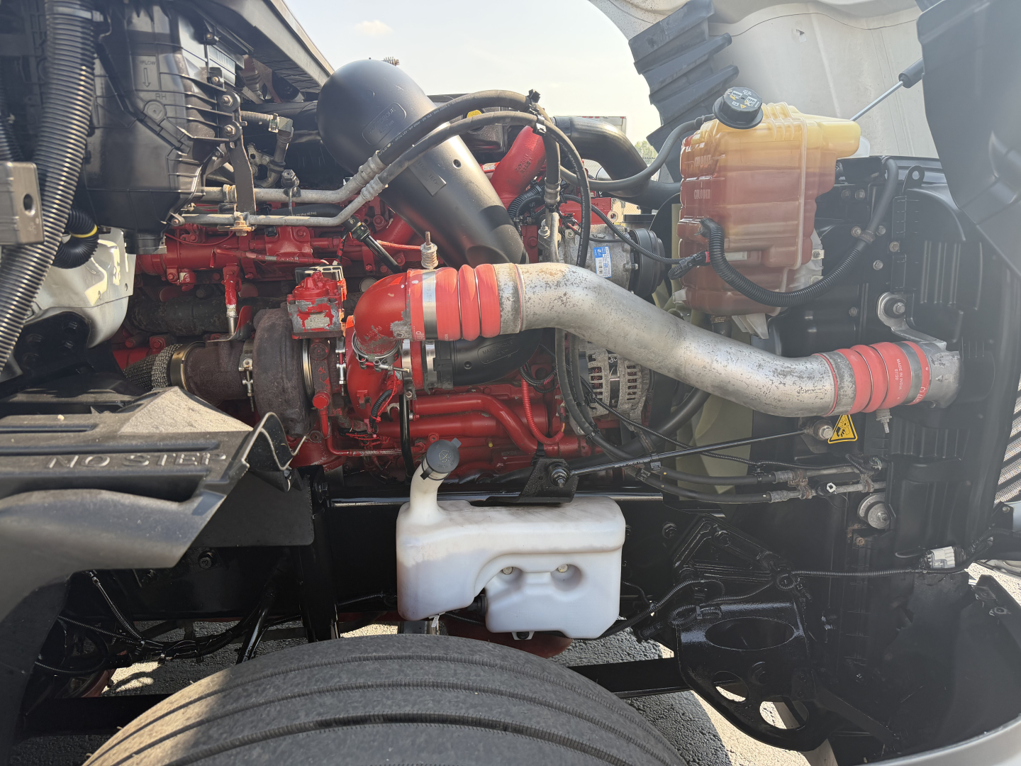 Truck engine bay with red engine, white coolant reservoir, and yellow fluid bottle. Visible hoses and metal components.
