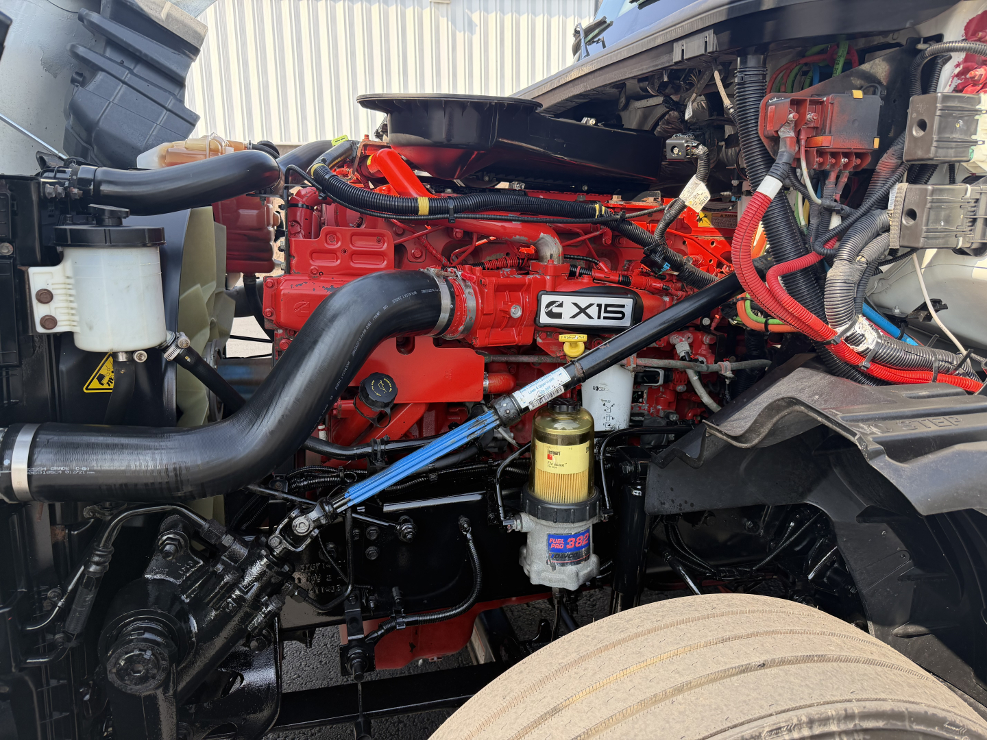Truck engine bay showing Cummins ISX-style red engine with black intake, yellow fuel filter, wiring harnesses, and radiator hoses.