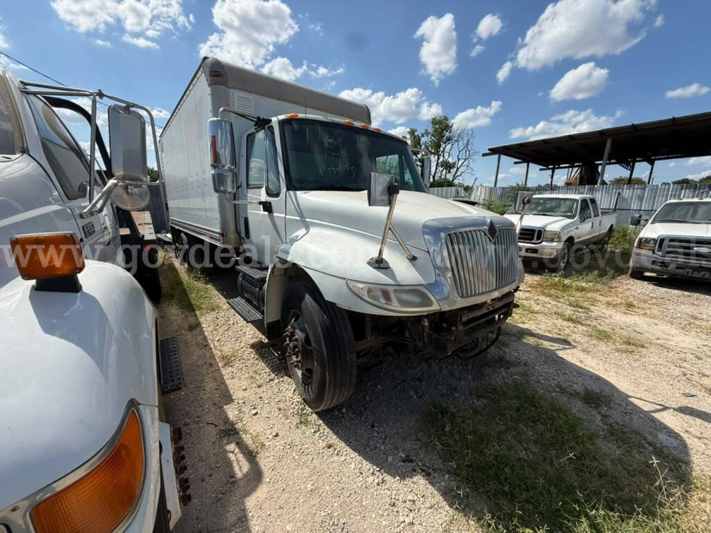 Truck cab exterior with box body, front view showing International logo, daytime, unused area around with other trucks in lot.