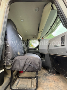 Truck interior cabin of a 2020 Mack Granite 64BR MHD showing driver seat and dashboard with Mack badge; vinyl seat cover, dirt floor, overhead ceiling, single cab layout.