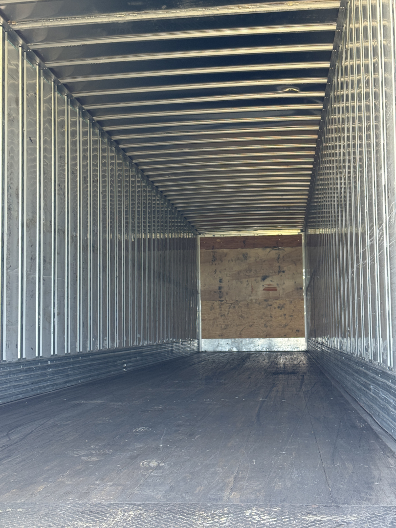 Interior of a utility trailer empty cargo space with metal walls and a wooden rear door, showing ribbed ceiling and floor boards.