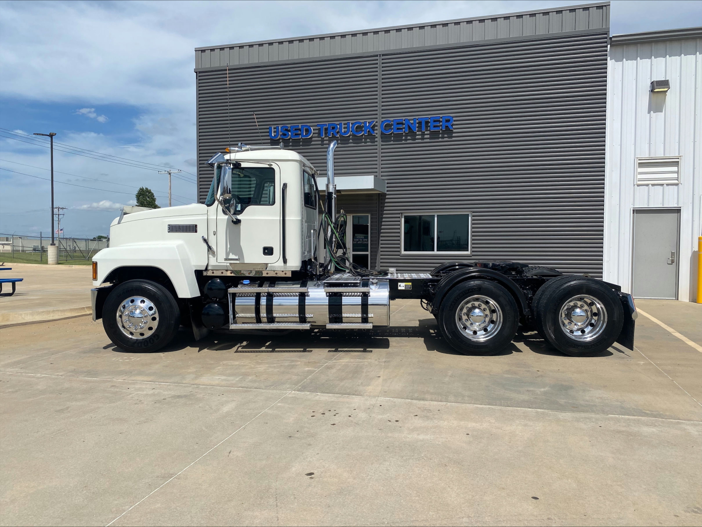 2021 Mack PI64T conventional daycab truck side profile showing white cab, tandem rear axles, and chrome fuel tanks in front of a used truck center building.
