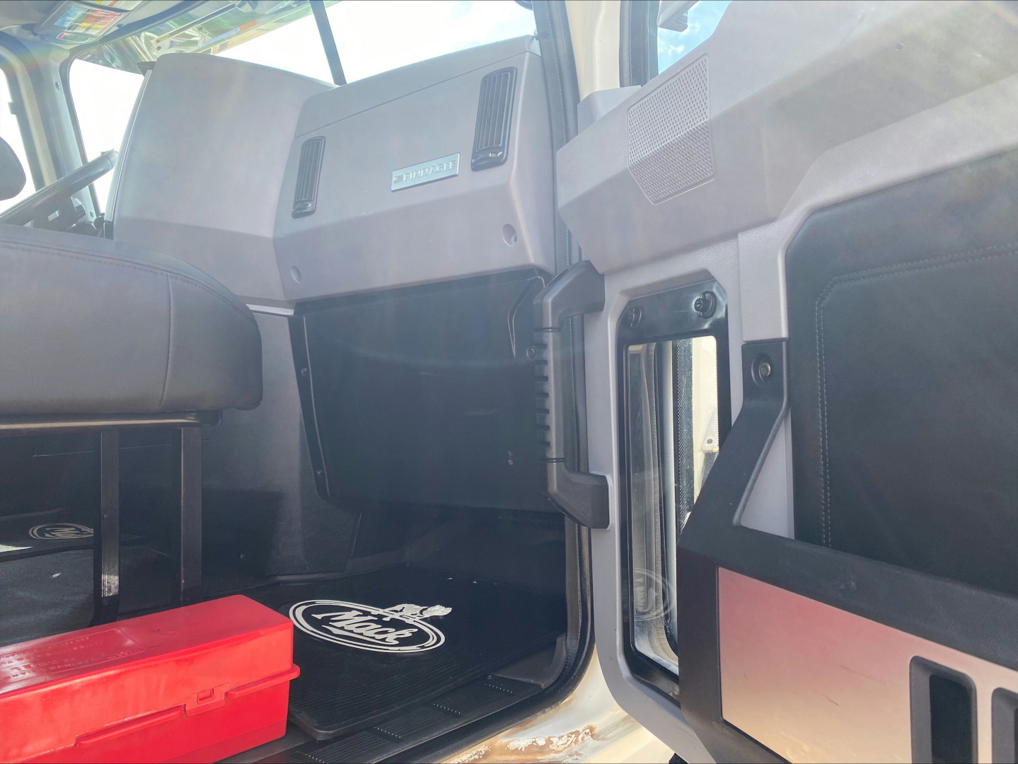 Cab interior of 2021 Mack PI64T showing driver footwell, open door, dashboard area with glovebox and vent, and a red tool box on the floor mat.