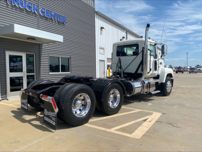 Conventional daycab truck in a dealership lot, Mack PI64T style, white cab with dual rear axles, chrome fuel tanks, large side mirrors, dealership building in background.
