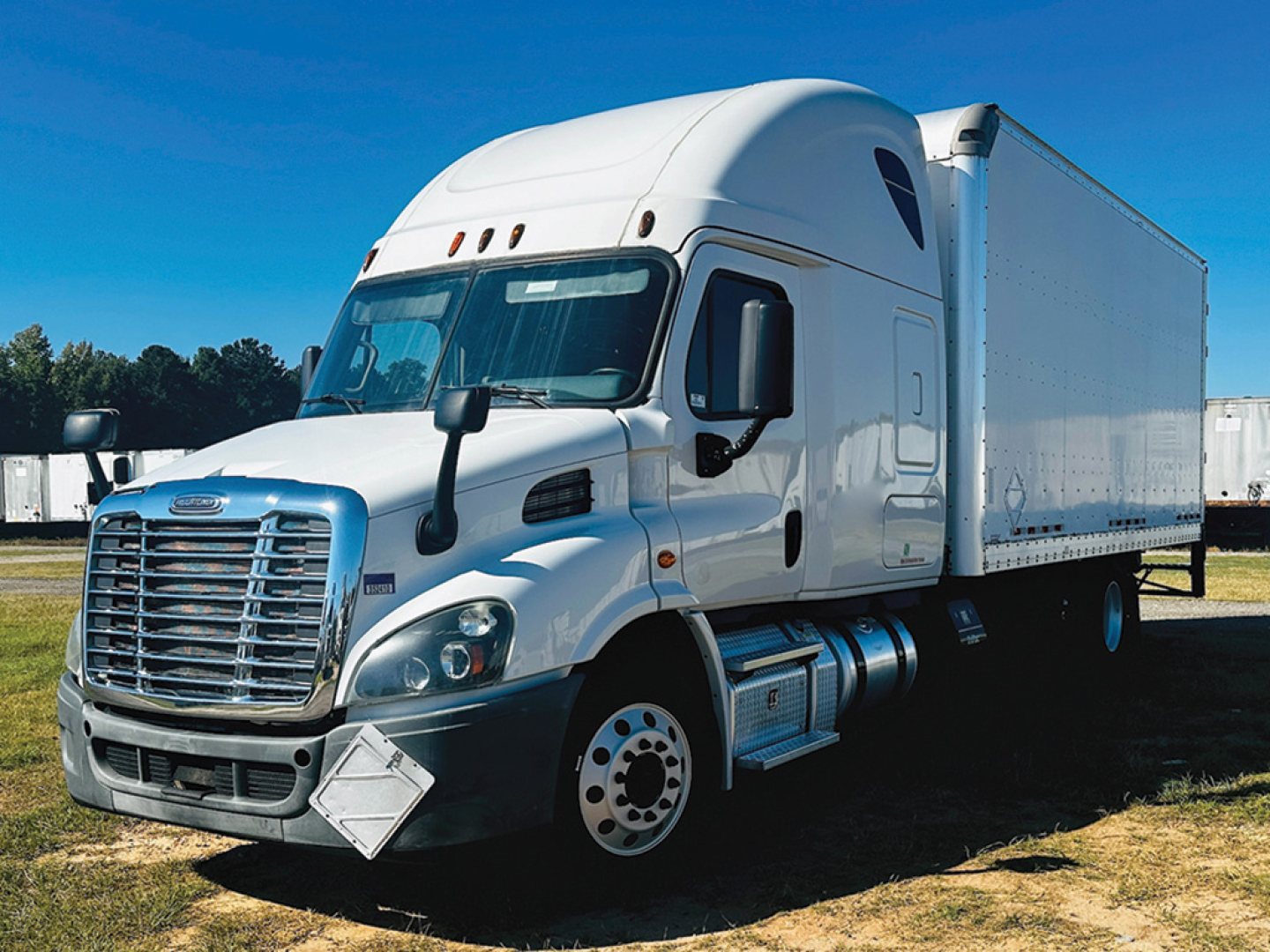 Truck in view showing a white Freightliner Cascadia 113 sleeper with a large box trailer attached, chrome grille, dual side fuel tanks, and aerodynamic cab.