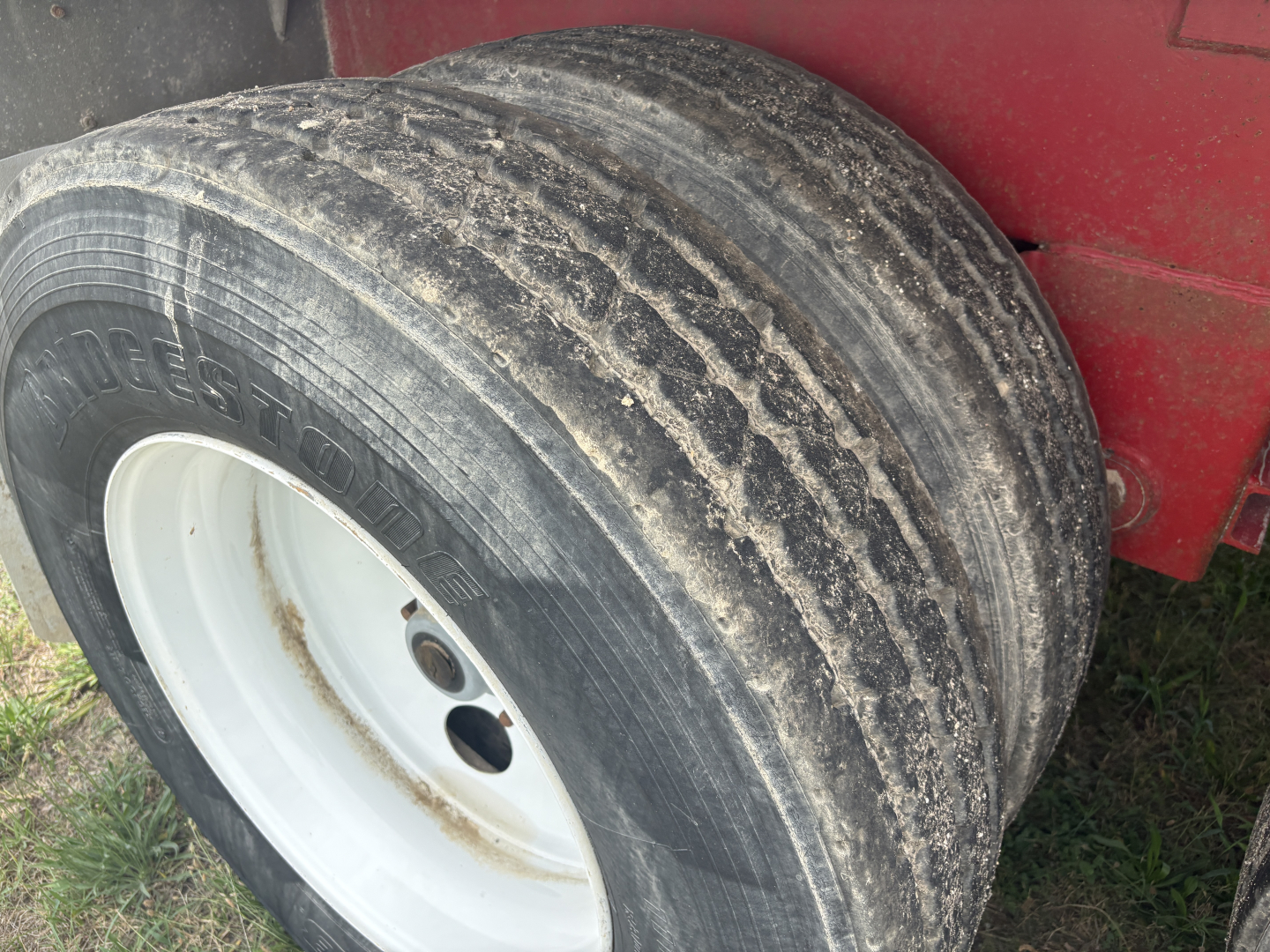 Front left dual-axle trailer wheel with worn tire treads and white rim; some dirt and grime on wheel well; partially obscured red trailer body.