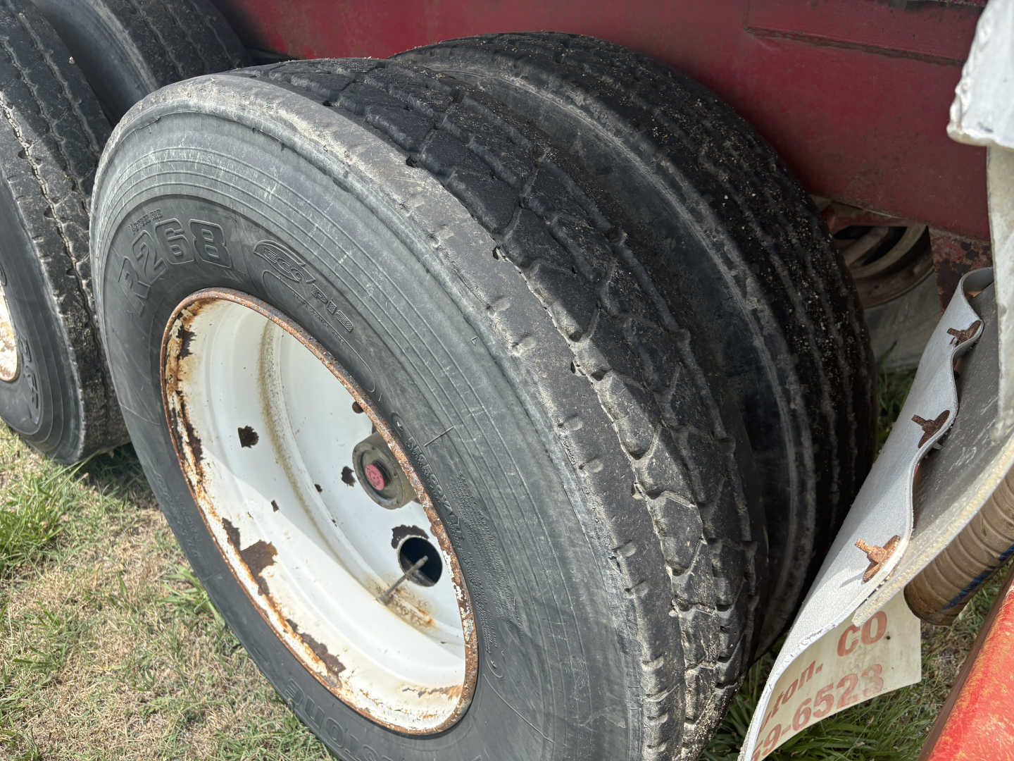 Trailer wheel with dual axle; close-up of black tire, white rim with rust spots, mud and grass in background.