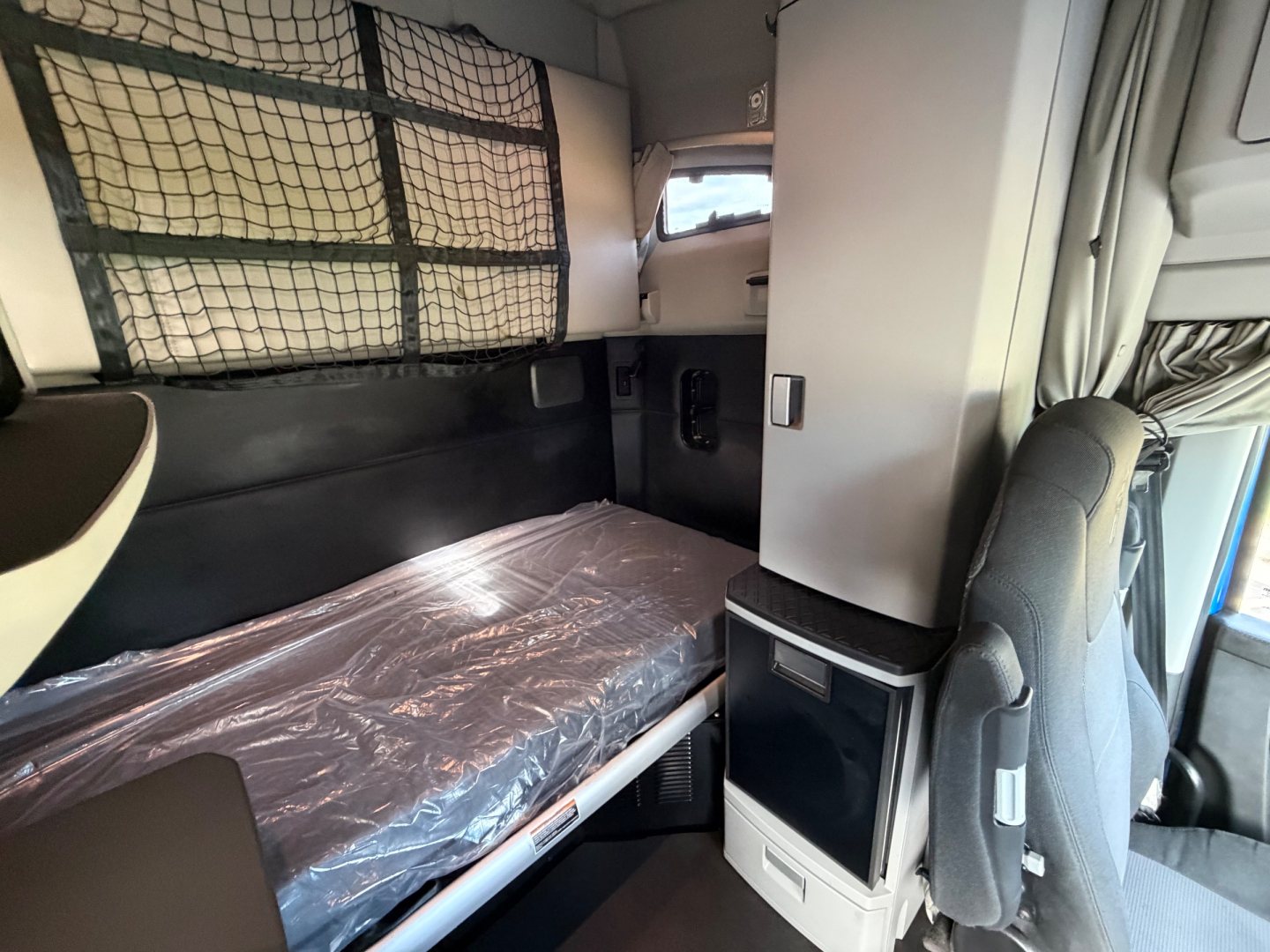 Kenworth T680 sleeper cab interior showing small bunk with plastic mattress, overhead cabinets, and a compact mini-fridge unit; grid barrier behind bunk.