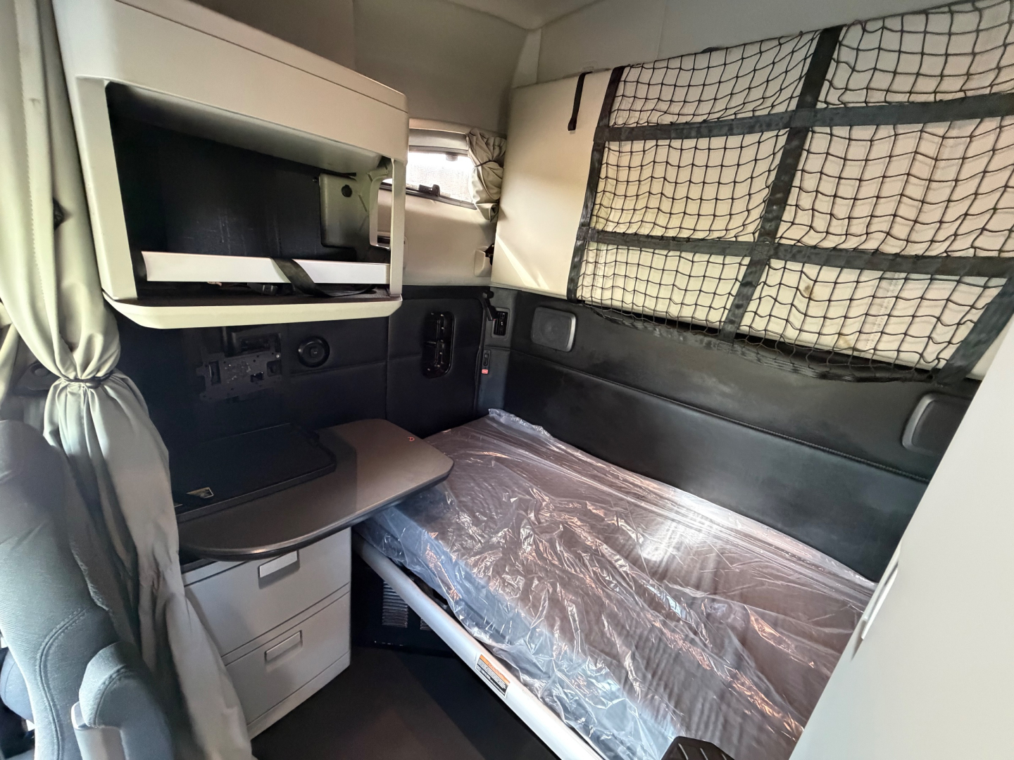 Kenworth T680 sleeper cab interior showing bed with plastic mattress cover, storage drawers, cabinet, and partition netting.