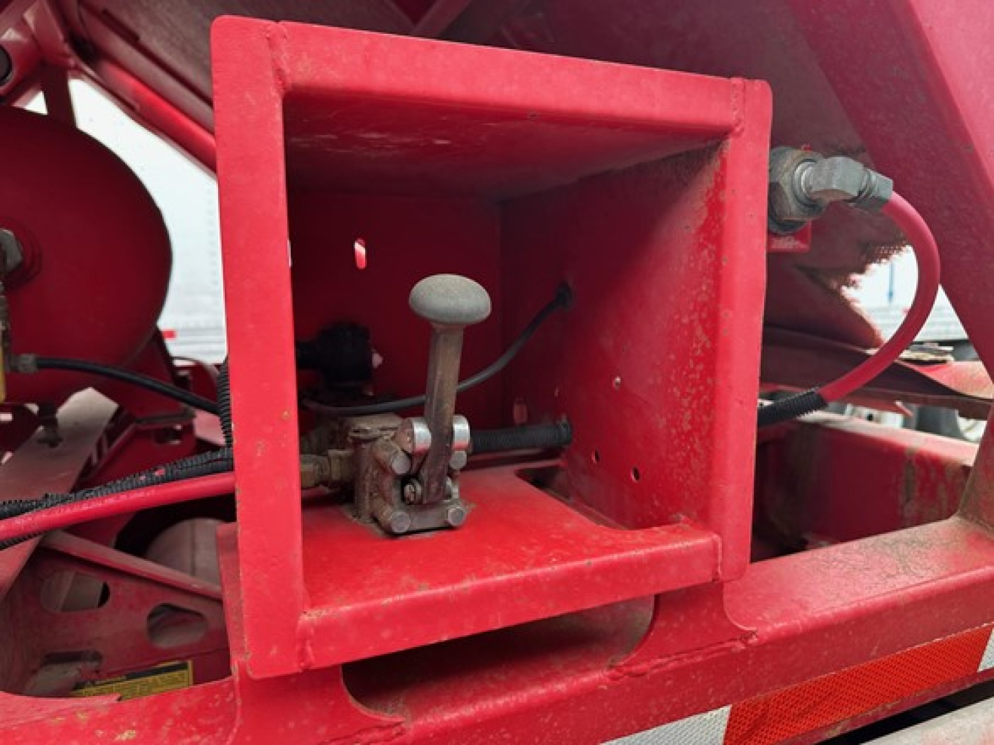 Red trailer hitch area with a square mounting box and a lever valve on a dump trailer. Visible hydraulic lines, frame, and bolts.