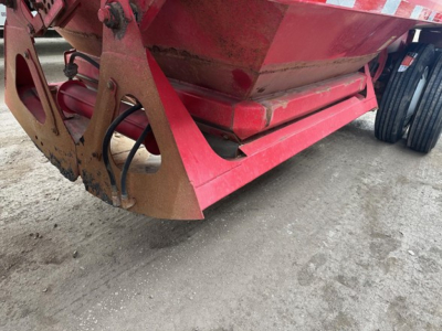 Front lower view of a red dump trailer attachment with hydraulic hoses and metal frame, showing chipped paint and dirt on the ground.
