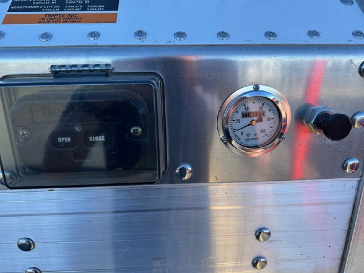 Hopper trailer control panel showing open/close switch and pressure gauge on stainless steel surface with bolts and label plate.