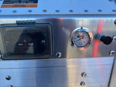 Hopper trailer control panel showing open/close switch and pressure gauge on stainless steel surface with bolts and label plate.