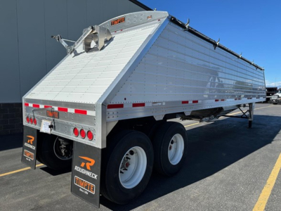 Timpte Roughneck hopper bottom trailer in aluminum with tailer design, rear dual axles, and reflective red/white stripes showing on side.