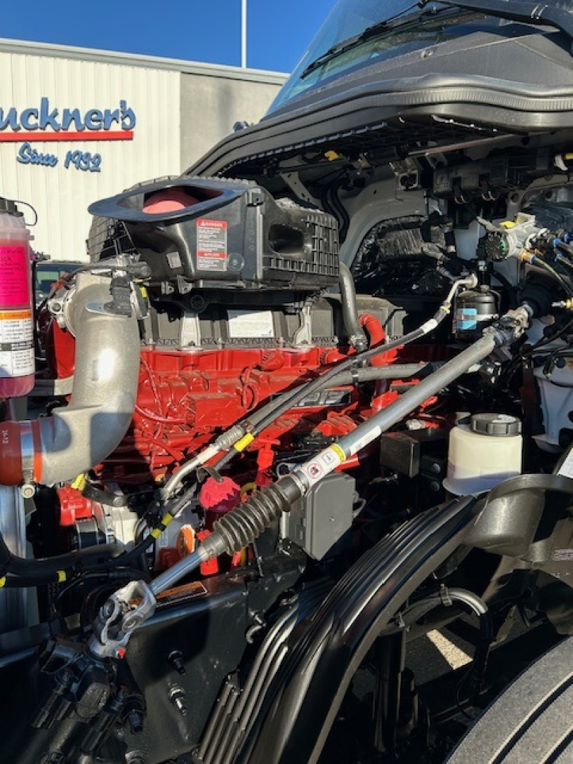 Truck engine bay of a new 2026 Mack PR64T64 showing red engine, air filter housing, hoses, belts, and hydraulic lines.