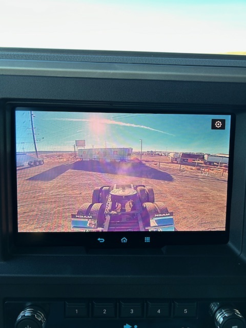 Truck cab monitor display showing tractor-semitrailer yard scene with chrome mirrors and dashboard buttons