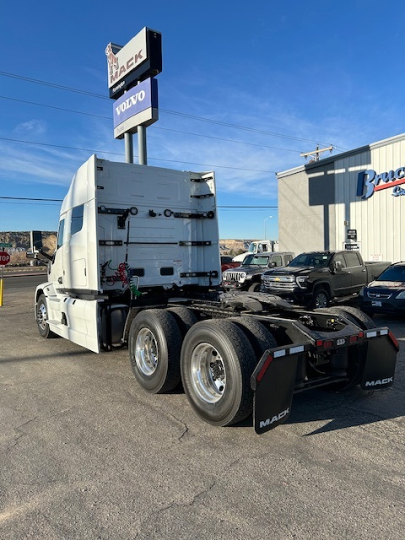 Truck cab and chassis of a 2026 Mack PR64T64 with rear triple axle, showing white cab, black dual rear axles, and mud flaps with 'MACk' branding. Visible dealership sign and surrounding lot.