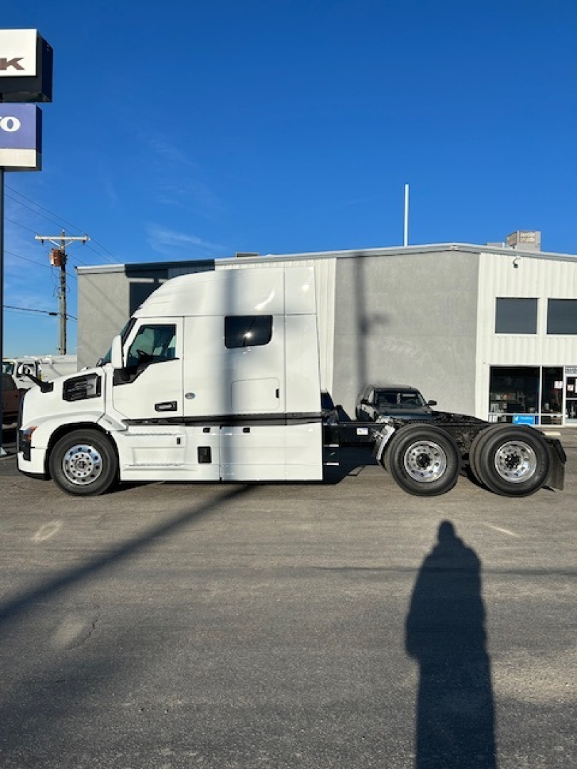 Truck side view of a new 2026 Mack PR64T64 conventional sleeper with three rear axles, white cab and black stripe, parked outside a building.