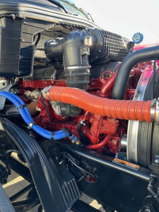 Truck engine bay view showing red engine, black radiator, orange coolant hose, blue hose clamps, and various hoses in a Mack PR64T64 truck.