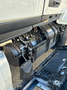 Engine area of a new Mack PR64T64 cab crop showing fuel/air tanks and engine bay components under a truck hood.