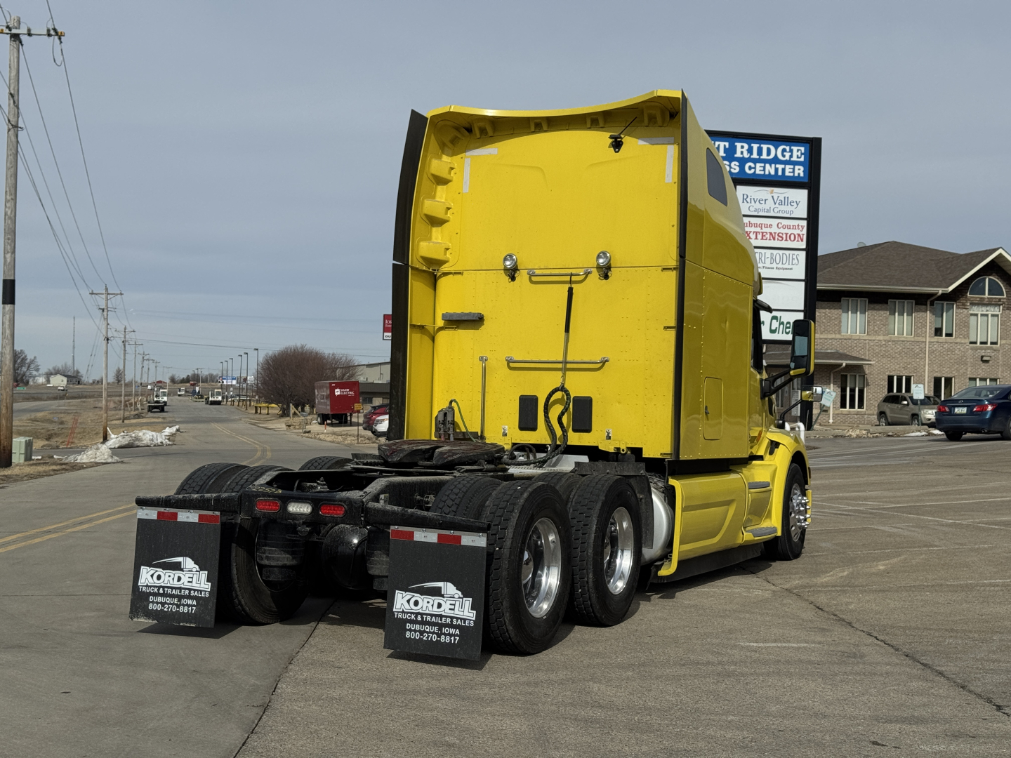 Peterbilt 579 truck in yard, rear view showing fifth wheel and trailer-axle area, yellow cab with black chassis and mud flaps with company branding.