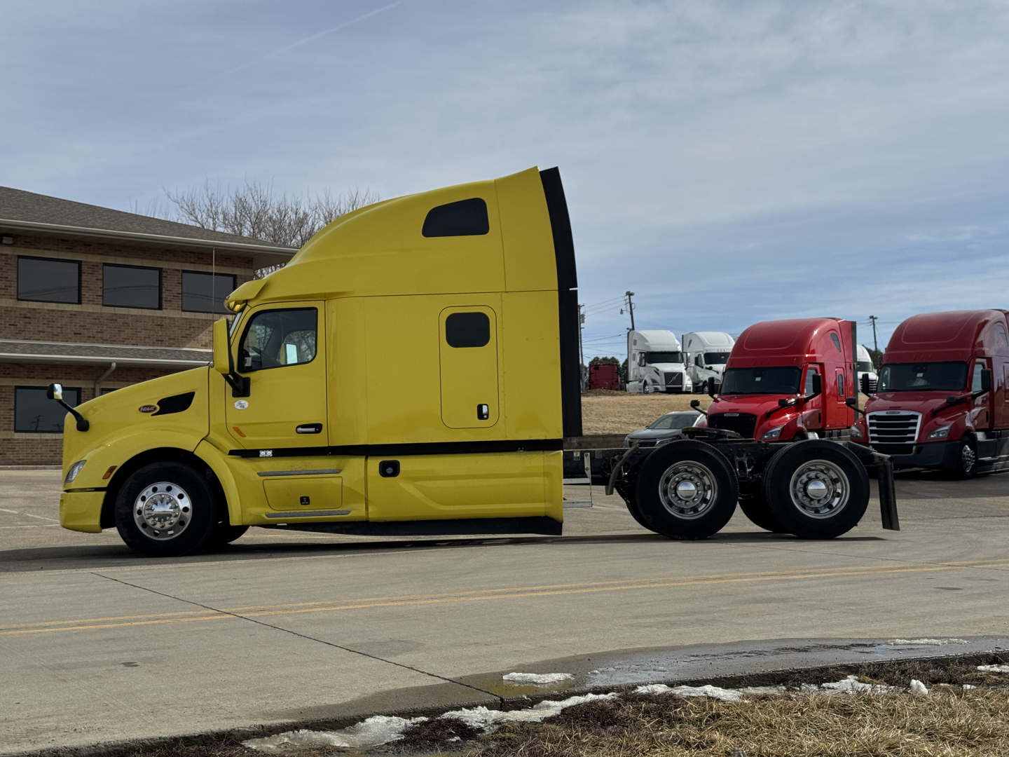 Yellow Peterbilt 579 truck cab in side profile with a black fifth wheel and red trucks in background.