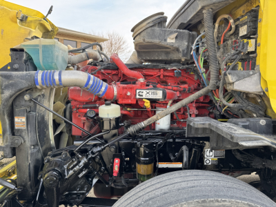Peterbilt 579 engine bay showing red engine with XIS badge, black hoses, wiring, and yellow cab frame; visible components include coolant reservoir, air intake, and various fluid lines.