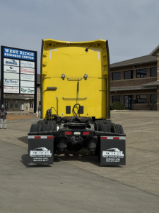 Peterbilt 579 rear view showing yellow sleeper cab, dual rear wheels, mud flaps, and dealership signage in background.