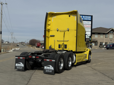 Peterbilt 579 truck in yard, rear view showing fifth wheel and trailer-axle area, yellow cab with black chassis and mud flaps with company branding.
