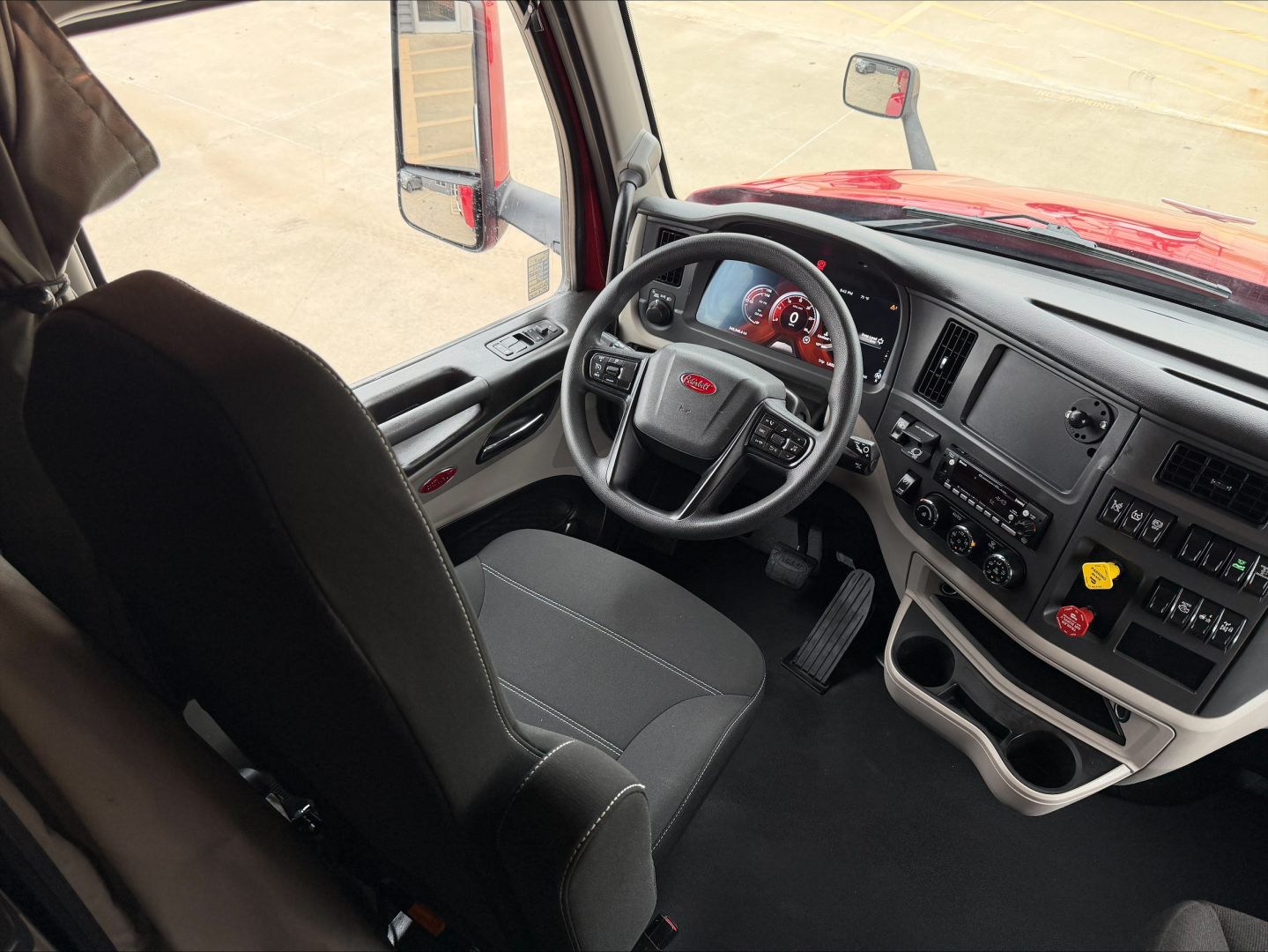 Peterbilt 579 truck cabin interior view showing steering wheel, dashboard, and driver seat from passenger side, red exterior visible, multiple control panels.