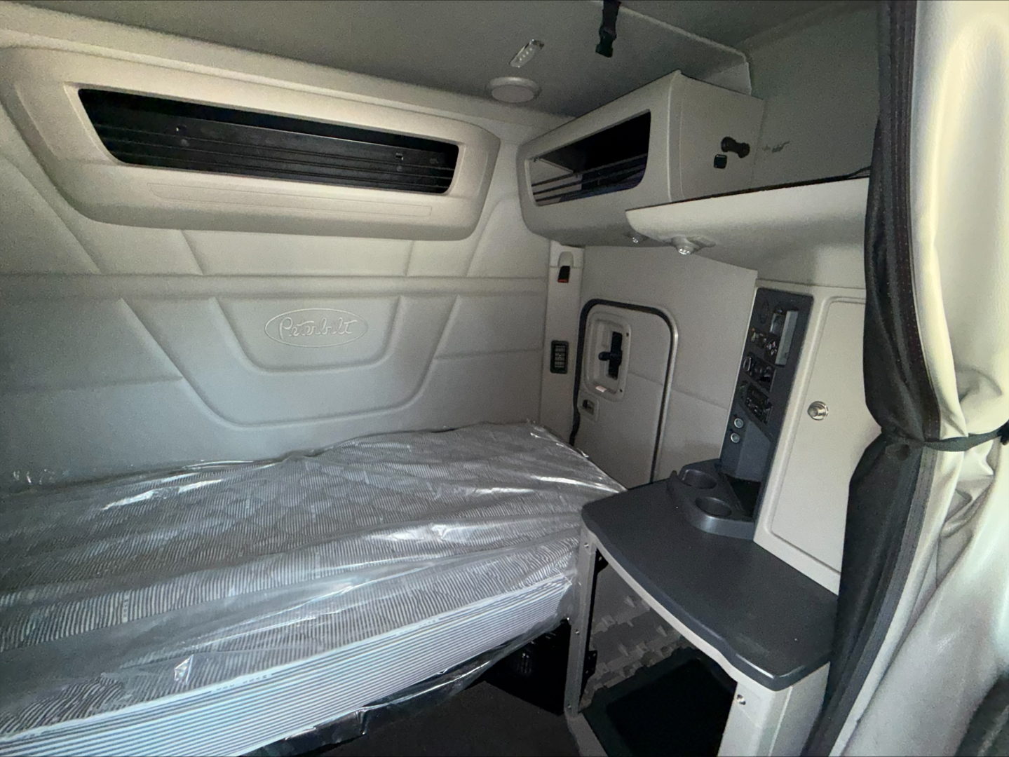 Peterbilt 579 sleeper cab interior showing bunk bed with plastic cover, overhead storage, and control panels on the passenger side.
