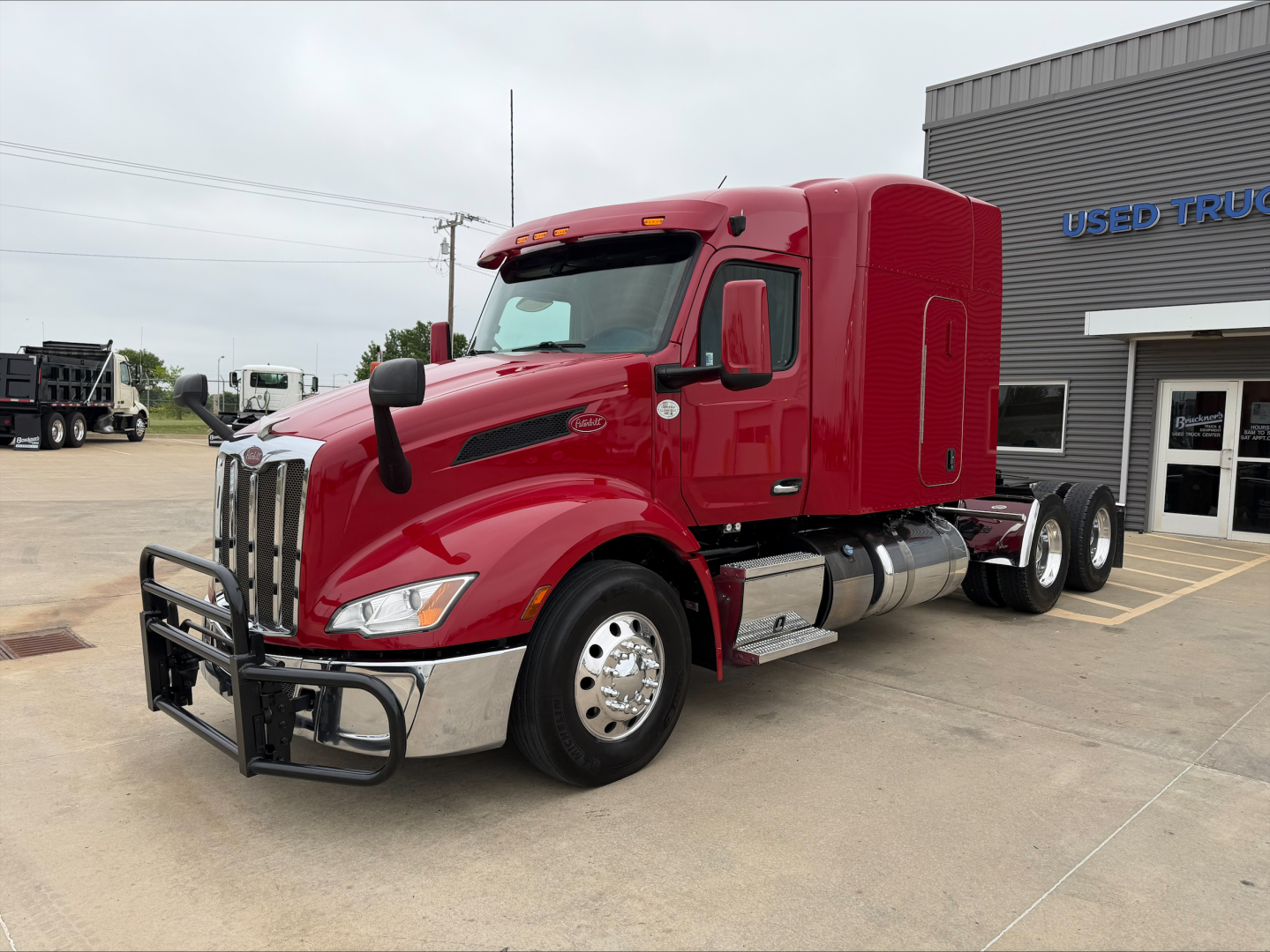 Peterbilt 579 truck in red, front left view with protective bumper, chrome fuel tanks, sleeper cab, dual rear axles, dealership lot.
