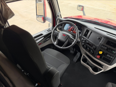 Peterbilt 579 truck cabin interior view showing steering wheel, dashboard, and driver seat from passenger side, red exterior visible, multiple control panels.
