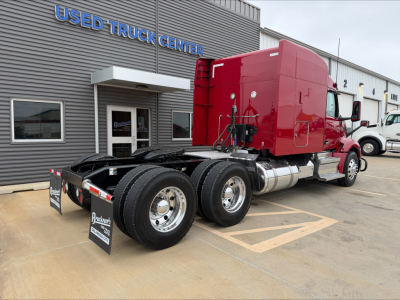 Peterbilt 579 tractor rear with red sleeper cab, view of chassis, dual rear axles, fuel tanks, and used truck center building in background.