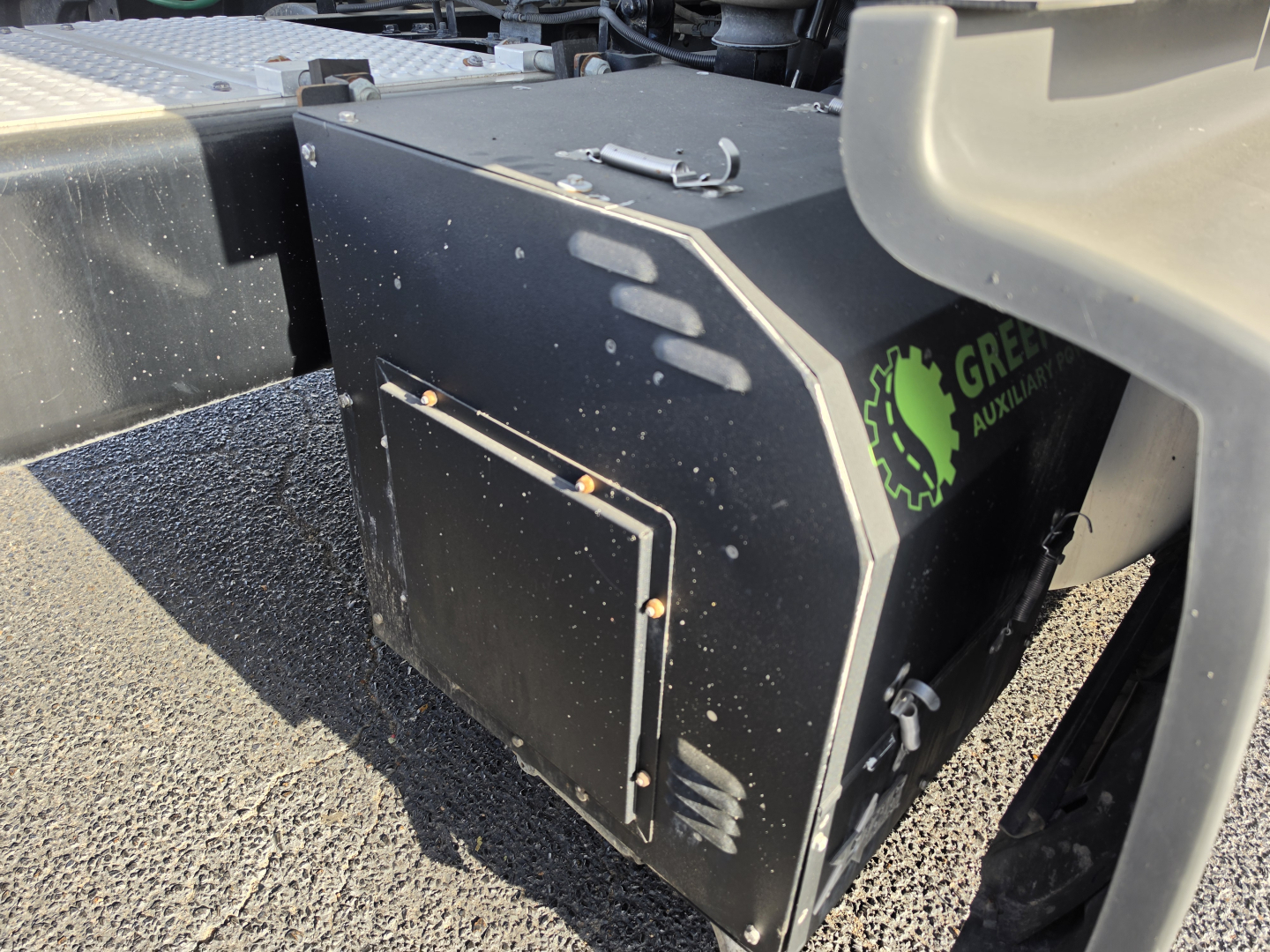 Truck component visible: black rectangular toolbox or housing with metal latches, mounted on chassis. Markings include a green circular logo with a gear and text 'GREEN' partially visible on the side. No other legible text.