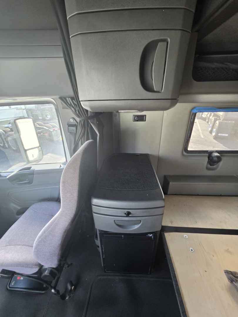 Interior cab area of a 2023 International LT625 sleeper, showing a storage drawer under a small cabinet, steering seat, and a plywood sleeping area; no visible text.