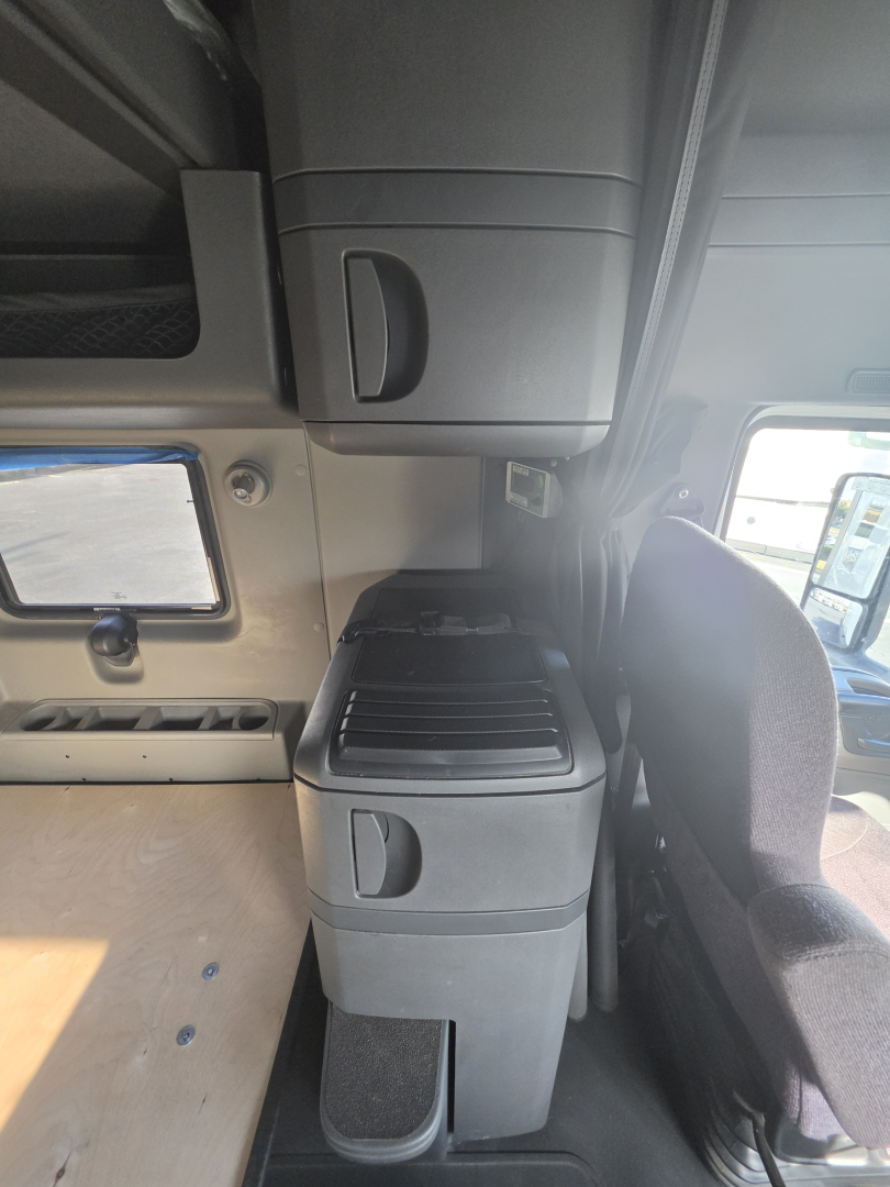 Cab interior showing overhead storage cubbies and a vertical stack with cup holder and drawers, driver seat on right side, window and door panel ahead.
