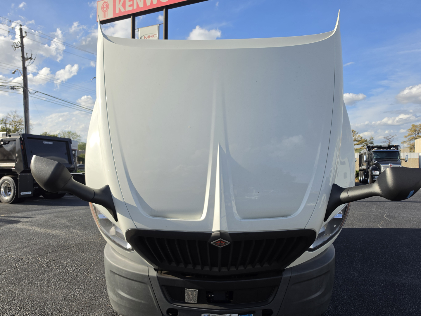 Truck hood and front grille of a 2023 International LT625 sleeper cab viewed head-on, with side mirrors and Kenworth dealership in background.