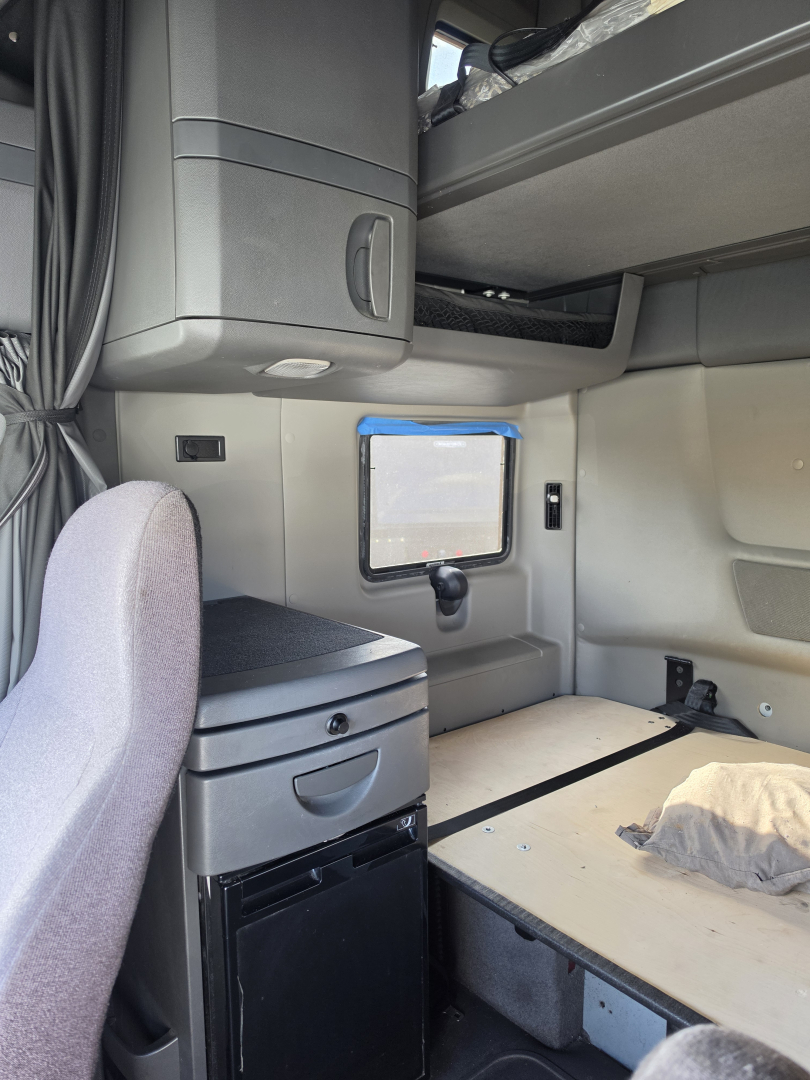 Truck sleeper cab interior showing side wall with small window, overhead bunk, storage cabinet, and compact fridge; bed area with light wood floor panel and a pillow.