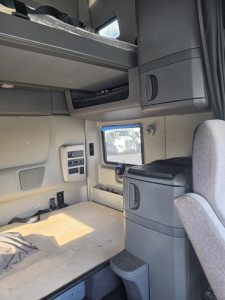 Cab interior of a used 2023 International LT625 sleeper truck showing lower bunk area, storage cabinets, and a small window; light gray interior with control panels on wall.
