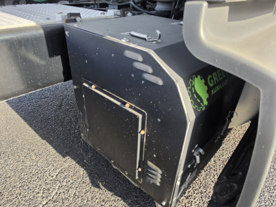  Truck component visible: black rectangular toolbox or housing with metal latches, mounted on chassis. Markings include a green circular logo with a gear and text 'GREEN' partially visible on the side. No other legible text.